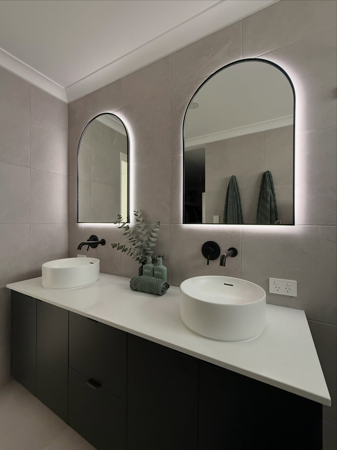 Two illuminated arch mirrors over white basins and a dark vanity