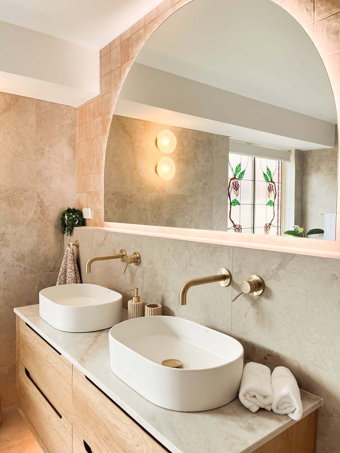 Warm stone and timber bathroom with white basins, gold tapware and a large curved mirror