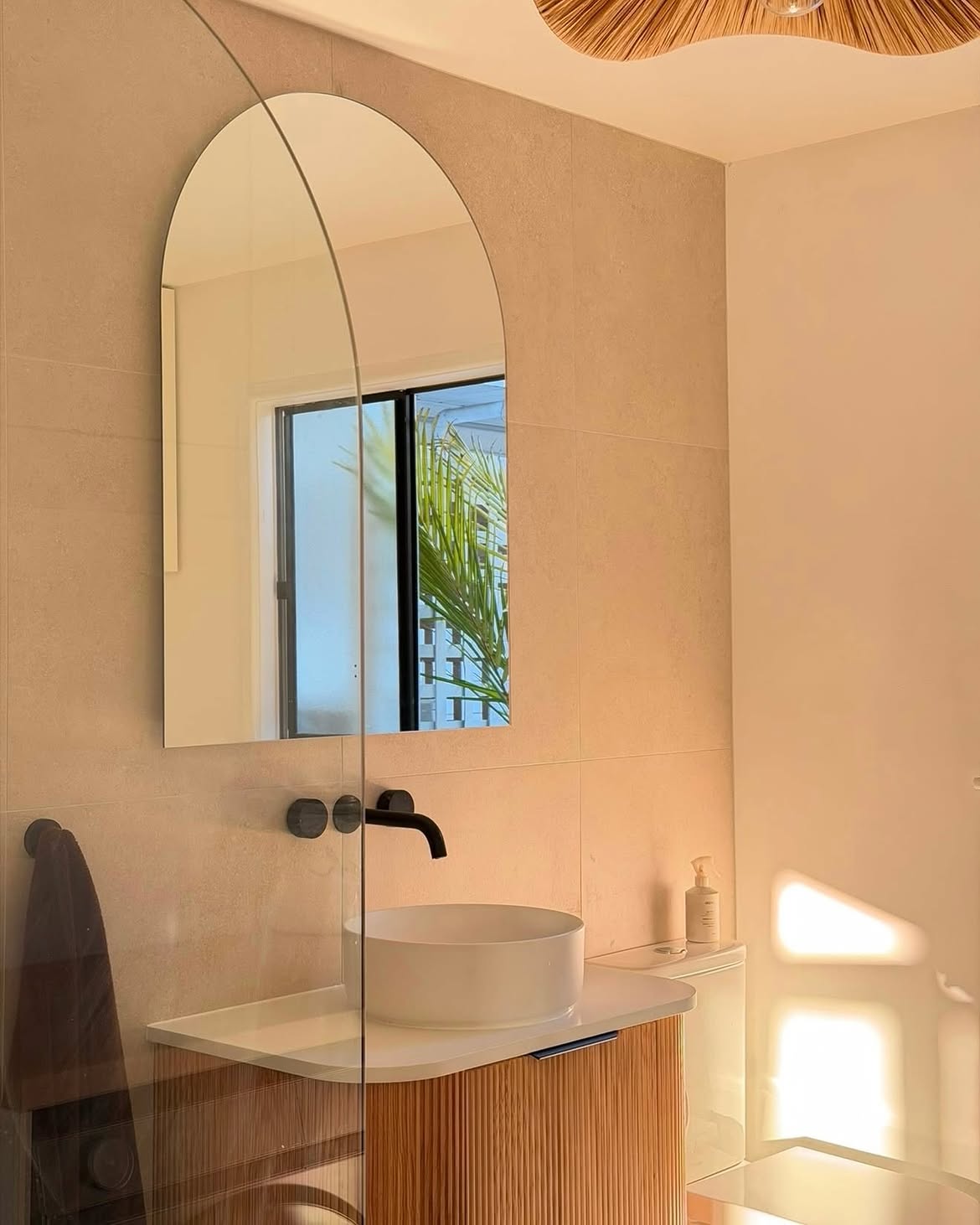 Sunlit bathroom with an arch mirror and curved timber vanity